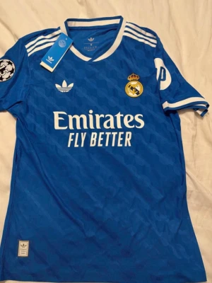 Real Madrid - Snygg blå Real Madrid matchtröja från Adidas med vita detaljer och klubbmärke på bröstet. Tröjan har korta ärmar, rund hals och tryckta loggor samt sponsortexten 'Emirates Fly Better' på framsidan. Tillverkad i lätt och ventilerande material.