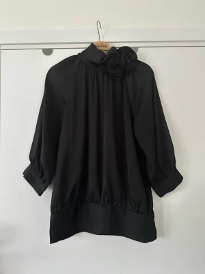 Svart VILA blus med blomdetaljer - storlek S - Snygg svart blus med hög krage och stor tygblomma på axeln. Blusen har trekvartsärm med breda manschetter och knappar. Tillverkad i ett glansigt, mjukt material som ger ett elegant intryck. Perfekt för dig som gillar stilrena plagg med en twist.