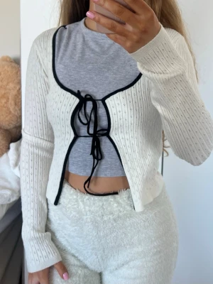 Kofta - Söt vit ribbad kofta med svarta kontrastkanter och knytband framtill. Koftan har lång ärm, croppad passform. Storlek XS från FB Sister (New Yorker) endast använd nån enstaka gång 💕