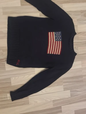 Mörkblå stickad tröja Polo Ralph Lauren - Mörkblå stickad tröja från Polo Ralph Lauren med amerikansk flagga framtill och RL-broderi vid nederkanten. Tröjan har lång ärm, ribbade muddar och klassisk rund hals.