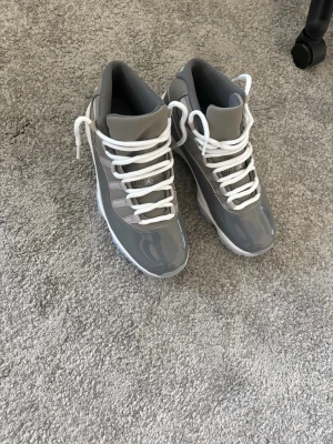 Gråa Air Jordan 11 sneakers - Snygga Air Jordan 11 sneakers i cool grå färg med vita skosnören och vit sula. Skorna har en blank finish i patentläder och meshdetaljer på ovandelen. Klassisk high-top modell som ger en sportig vibe och sticker ut i mängden. Vill sälja de så fort som möjligt, pris kan diskuteras:)
