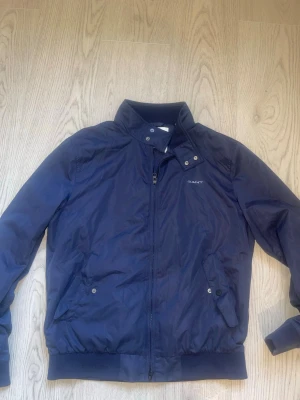 Marinblå vindjacka från GANT - Snygg marinblå vindjacka från GANT med hög krage och dragkedja framtill. Jackan har två snedställda fickor med knappstängning och ribbade muddar vid ärmslut och nederkant. Tillverkad i lätt och tåligt material, perfekt för blåsiga dagar.