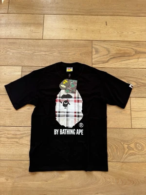 Svart BAPE t-shirt - Säljer en svart t-shirt från A Bathing Ape med ikonisk BAPE-logga framtill i rutigt och camouflage. T-shirten har rund hals, korta ärmar och tryckt text 'BY BATHING APE' nedtill. Perfekt för dig som gillar streetwear och vill sticka ut.