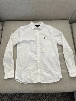 vit skjorta från Lyle & Scott - Klassisk vit skjorta från Lyle & Scott med gul logga på bröstfickan. Skjortan har lång ärm, knappar framtill och en enkel, stilren design. Perfekt till många olika looks och enkel att matcha.