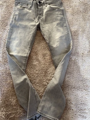 Gråa slim-fit jeans  - Säljer ett par gråa jeans i klassisk rak modell med fem fickor och bälteshällor. Jeansen har ljus tvätt och subtila slitningar framtill för en trendig look. Tillverkade i mjuk denim som ger en skön känsla och passar perfekt till streetwear-stil.