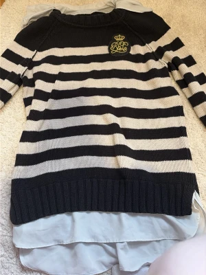 Randig stickad tröja från Ralph Lauren - Snygg stickad tröja från Lauren Ralph Lauren i marinblått och vitt med breda ränder. Tröjan har ett broderat RL-märke med krona på bröstet och rund hals. Långa ärmar och ribbstickade muddar. Perfekt för en klassisk och avslappnad stil.