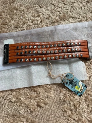 Humanity brunt läderarmband med nitar och text - Humanity. Coolt brunt armband i läder med flera rader, dekorerat med silvriga nitar och inspirerande texter ingraverade. Armbandet har ett brett spänne och detaljer som ger en edgy och personlig stil. Perfekt accessoar för dig som vill sticka ut.