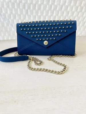 Blå axelväska med nitar Rebecca Minkoff - Snygg blå axelväska från Rebecca Minkoff i envelope-modell. Väskan har silverfärgade nitar på locket och en kedjerem i silver. Tillverkad i strukturerat konstläder som ger en cool och trendig look. Perfekt för dig som vill sticka ut med accessoarer.