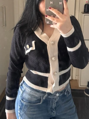 Svart kofta - Trendig svart kofta med vita detaljer och dekorativa pärlknappar. Koftan har v-ringning, långa ärmar och ribbade muddar. Snygg kontrast mellan svart och vitt, samt en fejkficka med knappdetalj på bröstet. Perfekt för en chic och stilren look.