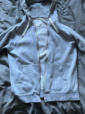 Ljusgrå zip hoodie med huva - Säljer en ljusgrå hoodie med dragkedja från 457. Hoodien har en klassisk huva med dragsko, två stora fickor framtill och ribbade muddar vid ärmslut och nederkant. Perfekt för en chill och avslappnad stil.