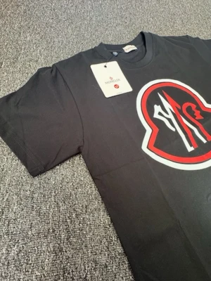 Svart Moncler t-shirt med stor logga - Svart t-shirt från Moncler med en stor, ikonisk logga i vitt och rött på bröstet. Klassisk rund halsringning och korta ärmar. Tillverkad i mjuk bomull som känns skön mot huden. Perfekt för dig som gillar streetwear och vill sticka ut. Helt ny aldrig använd 