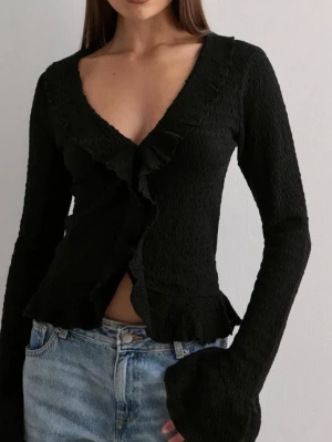 Svart volangblus med v-ringning - Svart blus med v-ringning och volangdetaljer längs framstycket och ärmsluten. Långärmad modell i strukturerad polyester med figurnära passform och lätt croppad känsla. Slutsåld!