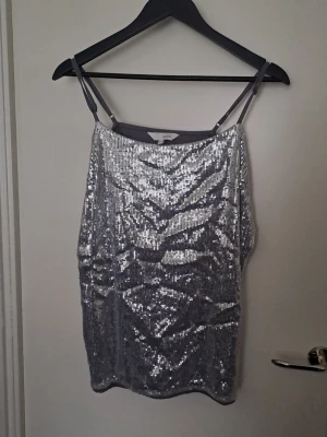 Glittrig silver linnetopp - Säljer en glittrig silverfärgad linnetopp med tunna axelband och paljetter på framsidan. Toppen har en rak passform och är perfekt för dig som vill sticka ut. Baksidan är i mjukt tyg utan paljetter för extra komfort. #Några paljetter saknas. Se bilder för övrigt skick 