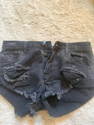 Svarta slitna jeansshorts One Teaspoon - Säljer ett par svarta, midwaist jeansshorts från One Teaspoon med råa kanter och slitna detaljer framtill och baktill. Shortsen har fem fickor, knappgylf och en avslappnad, loose passform. Perfekta för en edgy och trendig look.