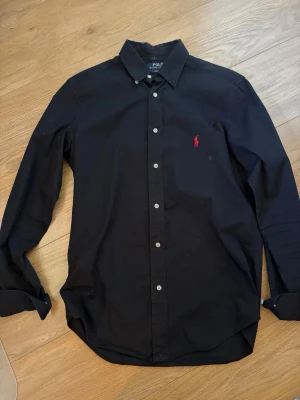 Ralph Lauren Skjorta  - Klassisk svart skjorta från Polo Ralph Lauren. Storlek S, passform Slim fit. Bra skick, ända defekterna är vid taggen i nacken som visas på bilden. Skriv vid frågor!