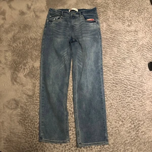 Levi's raka blå jeans - Klassiska blå raka jeans från Levi's med fem fickor och läderpatch bak i midjan. Jeansen har en avslappnad passform och tydliga sömmar. Tillverkade i slitstark denim med diskreta tvättade detaljer och små röda Levi's-taggar på baksidan. Jeansen är aldrig använda och har fortfarande kvar sina taggar.