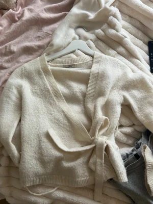 kofta ginatricot - Mysig krämvit stickad omlottkofta från Gina Tricot med knytband i midjan. Koftan har ribbstickade kanter och lång ärm, perfekt för lager på lager. Tillverkad i mjukt material som känns skönt mot huden.