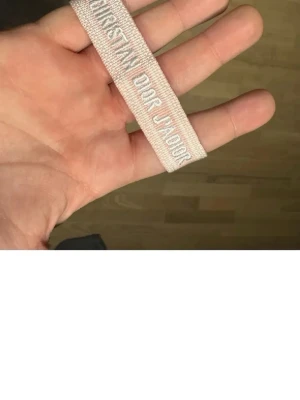 Christian Dior rosa tygarmband - Säljer ett ljusrosa tygarmband från Christian Dior med texten 'J'ADIOR' invävd i vitt. Armbandet har justerbara knytband och tofsar i ändarna. Perfekt accessoar för att lyfta din outfit med en exklusiv touch.