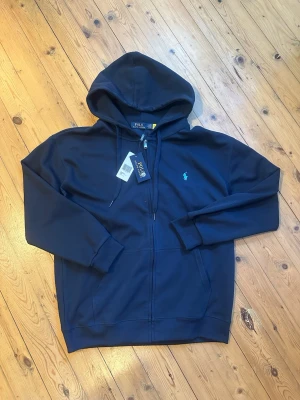 Marinblå hoodie från Polo Ralph Lauren - Snygg marinblå hoodie från Polo Ralph Lauren med dragkedja och klassisk huva. Ikoniska loggan i ljusblått broderad på bröstet. Mjuk bomullsblandning, ribbade muddar och fickor framtill. Perfekt för en chill och stilren look.