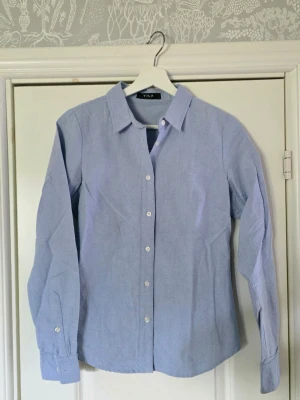 Ljusblå skjorta från Vila - Slim fit ljusblå skjorta från Vila med krage och vita knappar framtill. Skjortan har lång ärm och är tillverkad i bomull, perfekt för en clean och stilren look.