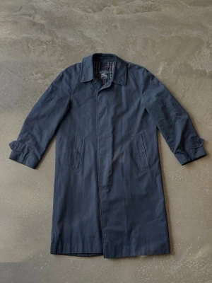 Vintage Burberrys Car Coat / Trenchcoat – Marinblå (46 REG) - Klassisk mörkblå trenchcoat från Burberry med rak passform och två snedställda fickor framtill. Insidan har det ikoniska rutiga fodret i grått, vitt och rött. Jackan har enkel krage och justerbara remmar vid ärmsluten. Perfekt för en stilren look.