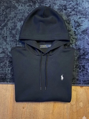 Svart hoodie från Polo Ralph Lauren - Snygg svart hoodie från Polo Ralph Lauren med klassisk huva och dragsnören. Ikoniska vita broderade loggan på bröstet. Tillverkad i mjukt bomullsmaterial som känns skönt mot huden. Perfekt för en avslappnad och stilren look.