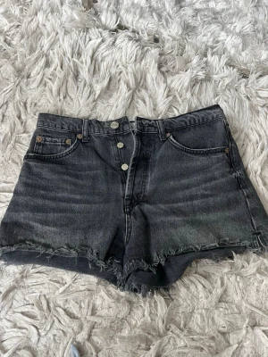 Svarta denimshorts - Säljer ett par svarta denimshorts med hög midja och rå, fransig kant. Shortsen har klassisk femficksdesign, knappar framtill. Perfekta för en avslappnad och trendig look.