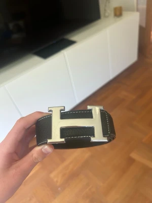  svart skärp med H-spänne - Säljer ett svart skärp med ett stort, ikoniskt H-spänne i silverfärgad metall. Skärpet är gjort i präglat skinn och har vita kontrastsömmar längs kanterna. Perfekt accessoar för att lyfta vilken outfit som helst.