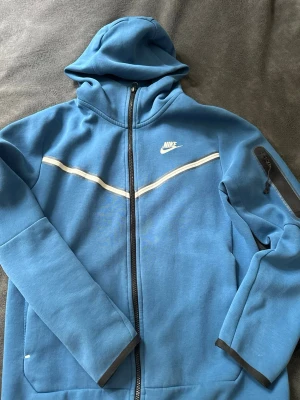 Blå zip hoodie från Nike - Snygg blå hoodie från Nike med dragkedja framtill och vit logga på bröstet. Hoodien har en vit rand över bröstet och en praktisk ficka med dragkedja på ärmen. Tillverkad i mjukt material och har huva för extra komfort.