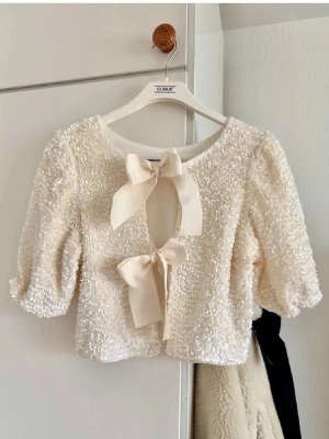 Vit paljettblus med rosetter - Supersöt vit blus med paljetter och två stora rosetter framtill. Blusen har puffiga trekvartsärmar och är croppad i modellen. Perfekt för dig som vill sticka ut lite extra med glittriga detaljer och romantisk vibe 💓 (Första bilden är inte min, lånad). Aldrig använd! Bara legat i garderoben tyvärr 