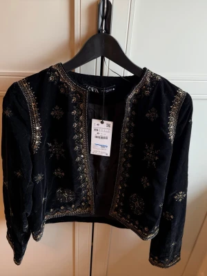 Svart sammetscardigan från Zara - Supersnygg svart sammetscardigan från Zara med detaljerade guldbroderier och öppen front. Kort modell med långa ärmar och rundad halsringning. Perfekt för att lyfta vilken outfit som helst med en lyxig touch. Helt ny.