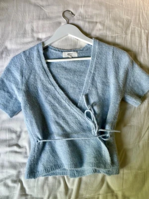 Ljusblå omlottopp  - Söt ljusblå omlottopp från Monki med korta ärmar och knytband i midjan. Tillverkad i ett mjukt, fluffigt material som ger en mysig känsla. Perfekt för dig som gillar en avslappnad men ändå trendig stil.