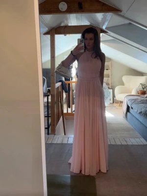 Ljusrosa långklänning med öppen rygg - Säljer en elegant ljusrosa långklänning med halterneck och öppen rygg. Klänningen har ett flytande, plisserat kjolparti och är ärmlös. Perfekt för student eller bal! Materialet känns lätt och svalt, troligen polyester.