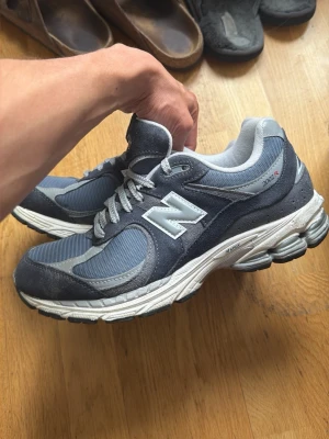 New Balance 2002R grå sneakers - Snygga New Balance 2002R sneakers i olika nyanser av grått och blått. Ovandelen är i mesh och mocka med vita detaljer och den klassiska N-loggan på sidan. Skorna har en chunky sula och reflekterande detaljer för extra stilpoäng.