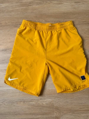Roger Federer Nike shorts - Säljer ett par gula Nike shorts med elastisk midja och RF-logga på vänster ben. Shortsen har en enkel design med vit Nike Swoosh på höger ben och är perfekta för sport eller chill. Materialet känns lätt och bekvämt.