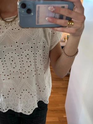 Vit broderad blus med spetsdetaljer - Superfin vit blus med broderade mönster och spetsdetaljer. Blusen är kortärmad och har en luftig känsla med vackra hålbroderier och vågig nederkant. Perfekt för en fräsch och somrig look.