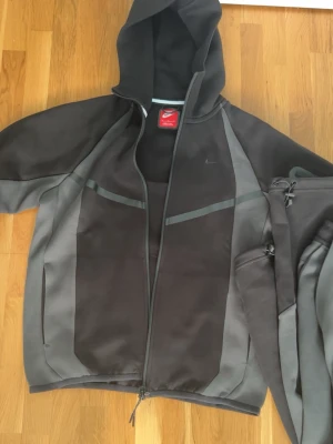 Svart och grå Nike hoodie med dragkedja - Nike hoodie i svart och grått med hel dragkedja framtill och huva. Snygga detaljer med mörkare paneler och diskret Nike-logga på bröstet. Tillverkad i mjukt och bekvämt material, perfekt för en sportig och avslappnad stil. Fickor på sidorna och ärmarna.