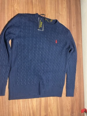 Marinblå kabelstickad tröja Polo Ralph Lauren - Kabelstickad marinblå tröja från Polo Ralph Lauren med rund halsringning och lång ärm. Klassisk röd broderad logga på bröstet. Tröjan har ribbade muddar och är tillverkad i mjukt material som passar perfekt till många outfits. Storlek S. Mysig och skön. 
