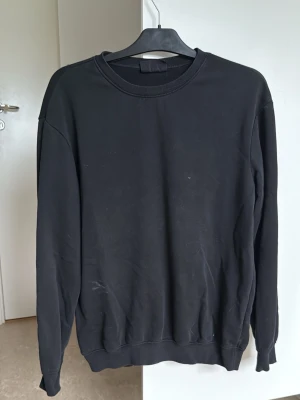 Svart basic sweatshirt - En enkel svart sweatshirt med rund hals och långa ärmar. Tröjan har ribbade muddar vid ärmslut och nederkant. Perfekt för en clean och avslappnad stil. Materialet ser ut att vara mjuk bomullsblandning.