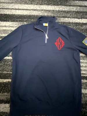 Efn quarterzip - Snygg mörkblå tröja med halv dragkedja och hög krage. På bröstet finns en broderad röd logga och på ärmen en patch med stjärnor. Perfekt till chill eller streetwear-stil. Materialet känns mjukt och bekvämt, och tröjan har långa ärmar. Epstein