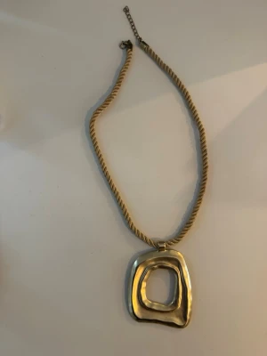 Trendigt halsband oanvänt - Unikt halsband med tjockt, tvinnat guldfärgat band och ett stort, abstrakt hänge i metall. Hänget har en organisk, oregelbunden form som ger en modern och trendig känsla. Perfekt för dig som gillar statement-smycken och vill sticka ut.