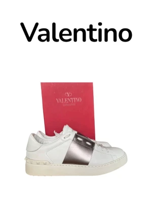 Valentino opens 40 - Snygga vita sneakers från Valentino med bred silverfärgad rem över mitten och ikoniska nitar på hälen. Skorna är i läder och har en klassisk låg siluett med vita snören och diskret logga på plösen. Perfekta för dig som vill ha en exklusiv touch till din outfit. Väldigt fint skick 9,5/10 som nya. Du får med allt og när du köper! Storlek 40