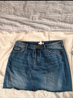 Blå jeanskjol med rå kant - Säljer en klassisk blå jeanskjol i kort modell med  fickor och rå, fransig kant nedtill. Kjolen har normal passform och stängs med knapp och dragkedja framtill. 