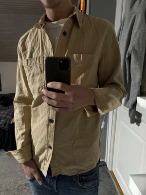 Beige overshirt - Stilren beige overshirt med klassisk krage och stora knappar framtill. Har en stor bröstficka på vänster sida och är tillverkad i ett lätt, strukturerat bomullstyg. Perfekt att bära öppen över en t-shirt för en avslappnad look.