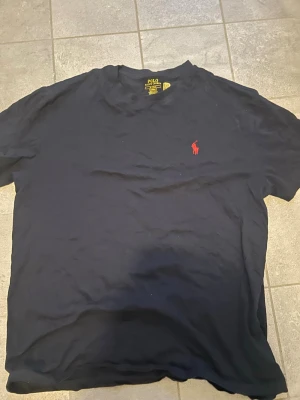 Svart t-shirt från Polo Ralph Lauren - Klassisk svart t-shirt från Polo Ralph Lauren med röd broderad logga på bröstet. Modellen är kortärmad och har en normal passform. Tillverkad i mjuk bomull som känns skön mot huden. Perfekt basplagg för en clean och stilren look.