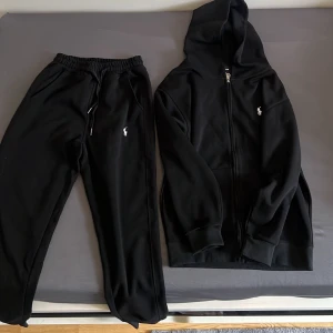 Ralph Lauren tracksuit  - Säljer en svart tracksuit från Polo Ralph Lauren med dragkedja framtill och klassisk huva. Tröjan har två fickor på magen och den ikoniska vita Polo-loggan broderad på bröstet. Tillverkad i mjukt bomullsmaterial som är skönt att ha på sig.