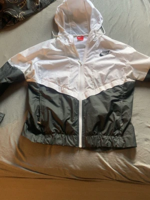 Nike svartvit vindjacka med huva - Nike vindjacka i svart och vitt med dragkedja framtill och huva. Jackan har elastisk nederkant och är tillverkad i lätt, vindtätt material. Klassisk Nike-logga på bröstet. Perfekt för en sportig och avslappnad stil.