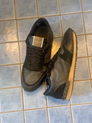 Svarta sneakers från Valentino - Säljer ett par svarta sneakers från Valentino med coola kamouflagemönster och detaljer i mocka och läder. Skorna har en beige sula med nitar på hälen och svarta skosnören. Perfekta för dig som gillar streetstyle och exklusiva märken.