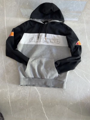 Ellesse hoodie i svart och grå - Snygg hoodie från Ellesse i svart, vitt och grått med stor logga över bröstet. Huva med dragsko och känguruficka framtill. Färgblockad design och broderade Ellesse-loggor på ärmarna. Perfekt för en avslappnad streetwear-look.
