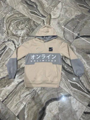 Beige hoodie med grå detaljer - Snygg beige hoodie med gråa partier på ärmar och bröst. Stor vit text med japanska tecken och 'UNLIMITED' framtill. Huva med dragsko och magficka. Perfekt för dig som gillar streetwear och vill sticka ut med cool design.